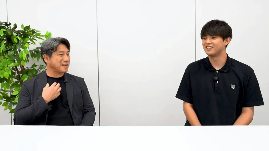白い背景の明るい部屋で、二人の男性が座って会話をしている様子