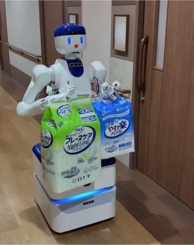 介護施設のような廊下で、人型ロボットが尿とりパッドと大人用パンツ型おむつの袋を両手に持ち、運搬している様子です。介護現場でのロボット活用を示唆する一枚。