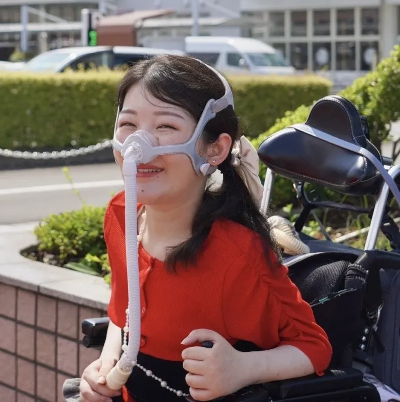 車椅子に座り、呼吸器マスクを装着した若い女性が屋外で明るく微笑んでいます。医療機器を使用しながらも、前向きに日常を送る様子が伝わる一枚です。