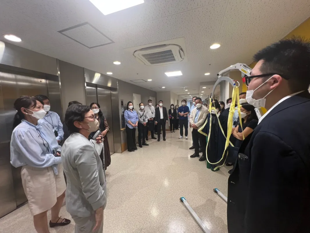 マスクを着用した人々が、エレベーターのあるモダンな屋内の廊下に集まっています。医療機器のようなものも見えることから、会議やプレゼンテーション、あるいは施設見学が行われている様子です。