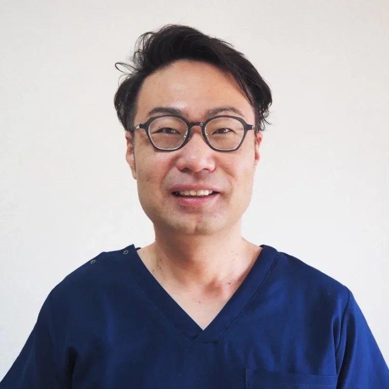 青いスクラブと眼鏡をかけたアジア人男性が、白い背景の前でカメラに向かって穏やかに微笑んでいるポートレート写真です。医療従事者や専門家のような印象を与えます。