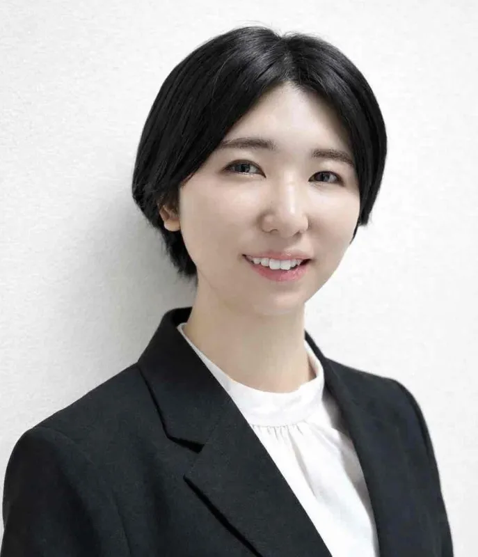 白の背景の前で、黒いショートヘアの若い女性が、黒いジャケットと白いブラウスを着用し、穏やかな笑顔を見せている上半身のポートレートです。プロフェッショナルで清潔感のある印象を与えます。