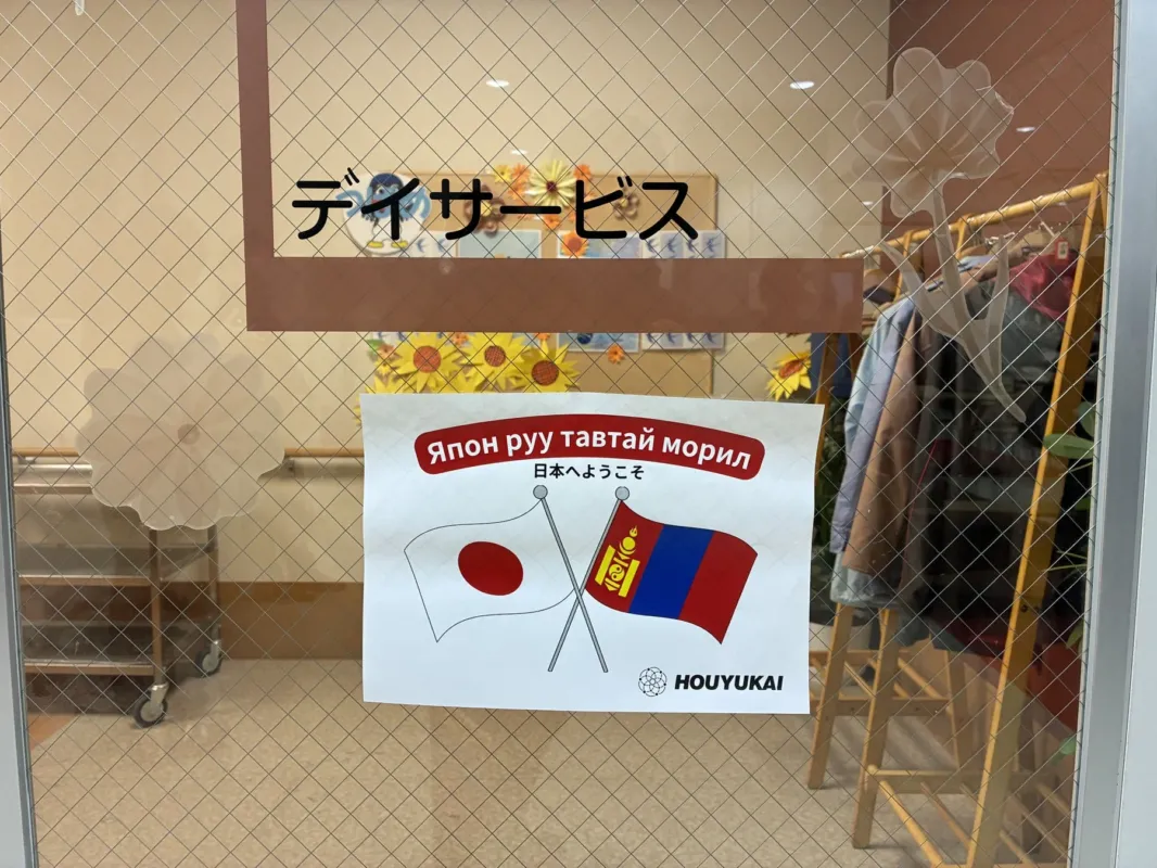 デイサービス施設のガラス戸に貼られたポスターの画像です。ポスターには日本とモンゴルの国旗が交差して描かれ、「日本へようこそ」とモンゴル語と日本語で書かれています。施設名「HOUYUKAI」も確認できます。モンゴルからの利用者や訪問者を歓迎している様子がうかがえます。
