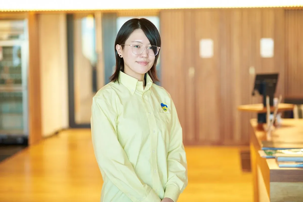 明るい木目調の室内に立つ、黄色いシャツと眼鏡をかけた若い女性のポートレート