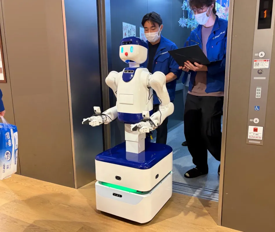 エレベーターの前に立つ人型サービスロボットと、それを見守るマスク姿の2人の開発者。ロボットは移動型の台座を持ち、一人の開発者はタブレットを操作している。